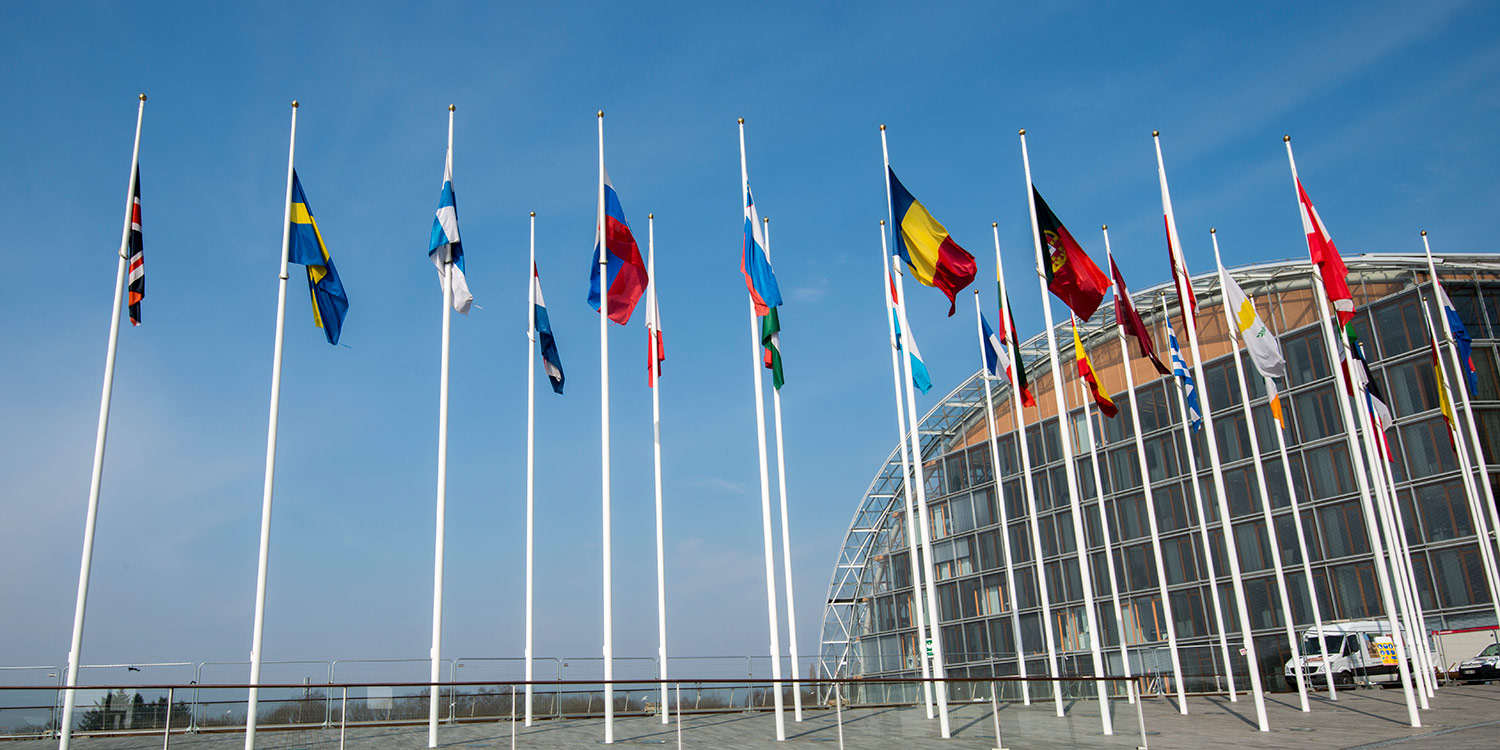 Looking upwards, the picture shows flagposts with EU country flags and an office building behind them.