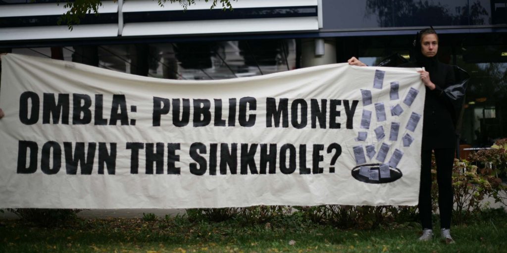 A young woman holding a banner that reads "Ombla: public money down the sinkhole?"