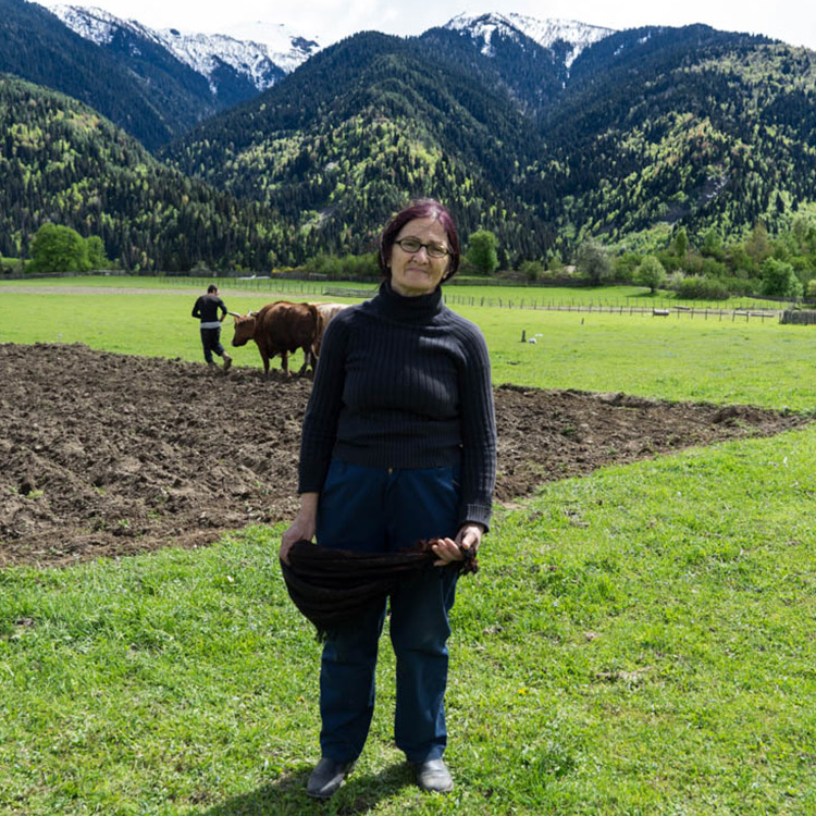 A woman looking into the camera is standing on a field with mountains in the background.